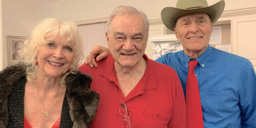 three friends standing together, woman on left in red dress, man in middle in red shirt, man on right in blue shirt and red tie and cowboy hat